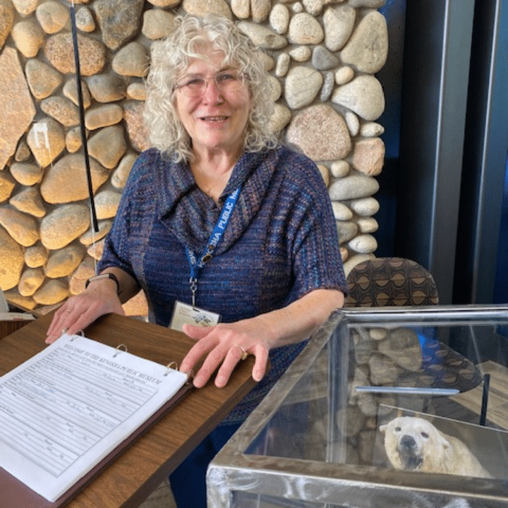 A person at a podium with a binder, a stone wall behind, next to a donation box.