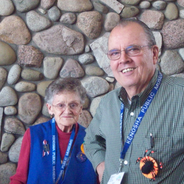 Two people smiling in front of a stone wall, wearing lanyards.