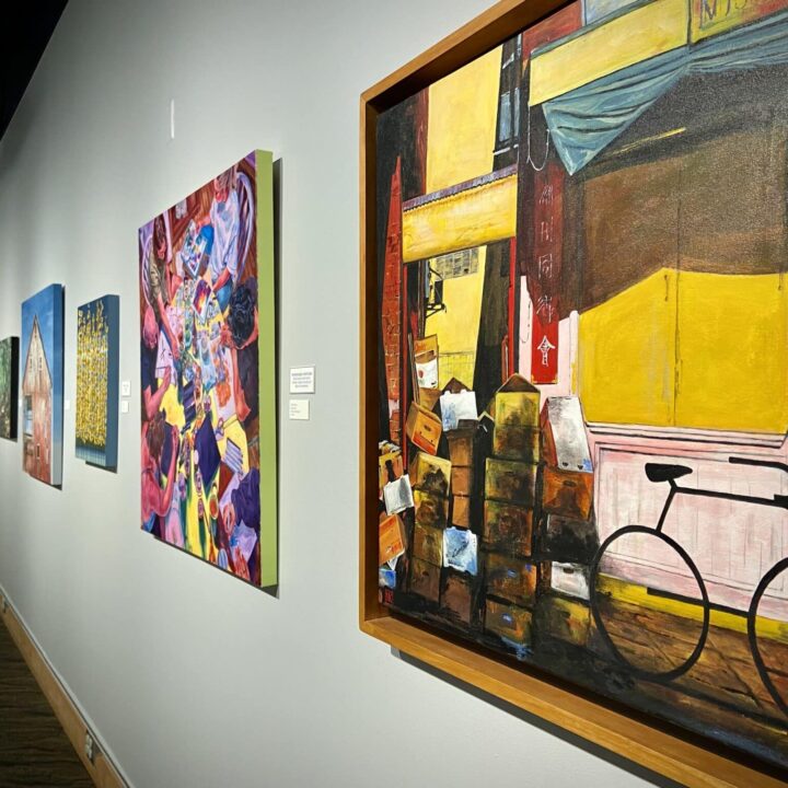 A gallery wall with colorful paintings, including a large street scene with a bicycle and boxes in the foreground.