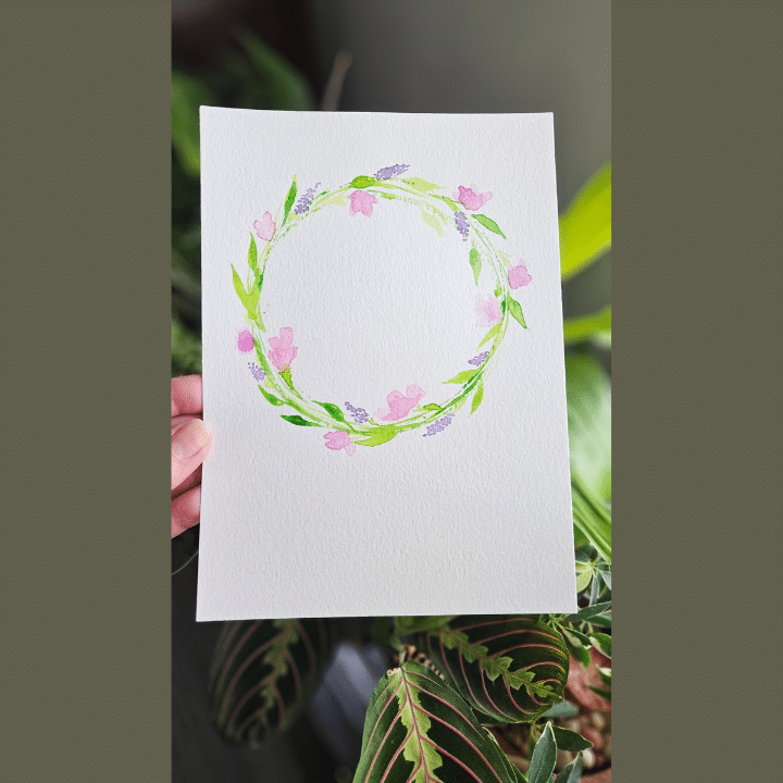 PaintSip_Wreath_720x720 A watercolor painting of a circular wreath with green leaves and pink and purple flowers on white paper, held by a hand.