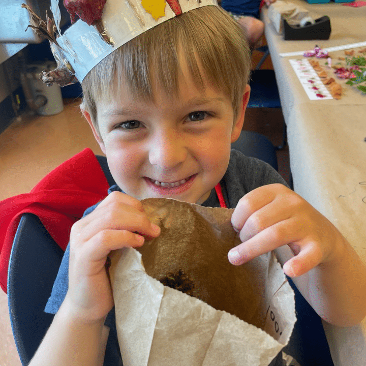 Child wearing a leaf-adorned crown, holding an open paper bag while sitting at a craft table.