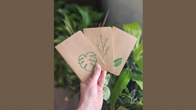 Hand holding three brown notebooks with embroidered plant designs against a leafy background.