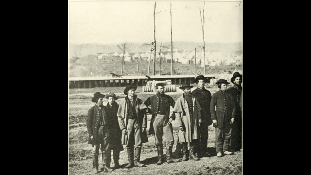 A group of nine soldiers in uniform standing on dirt with a military camp and tents in the background.